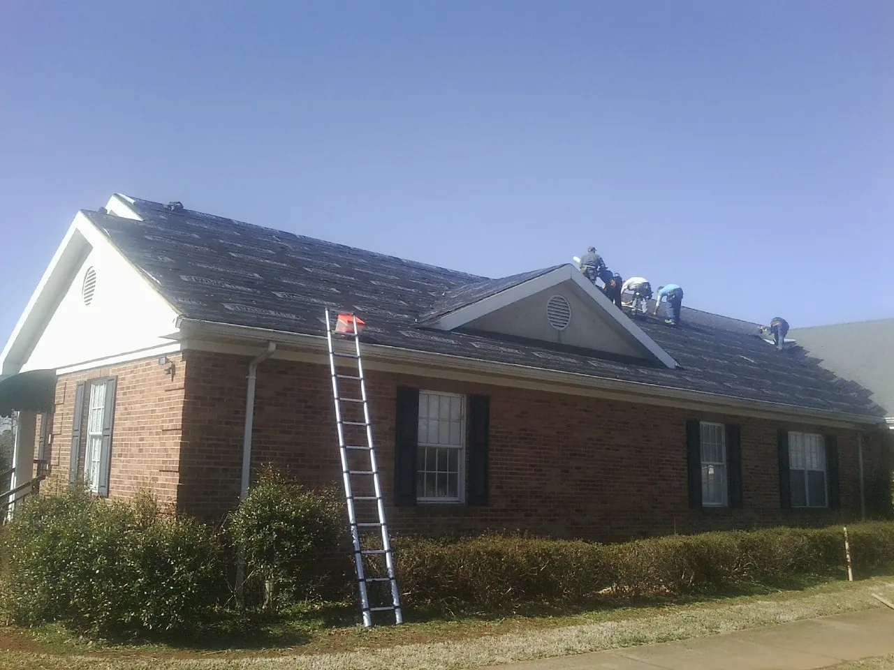 Crew tearing shingles off a brick ranch roof with a ladder set — Metal Roof Repair work by our crew in Philipsburg