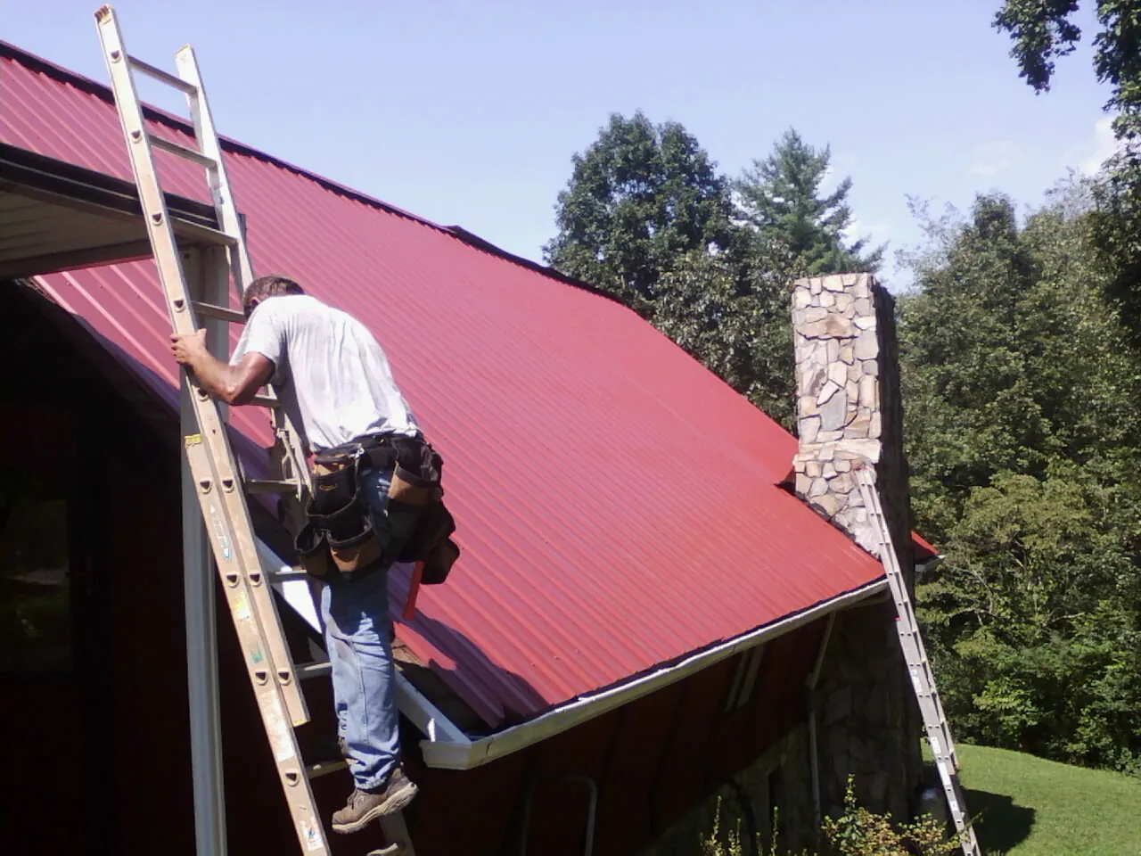 Roofer on a ladder beside a red metal roof and stone chimney — Soffit & Fascia Repair by Roofing Brothers in Philipsburg